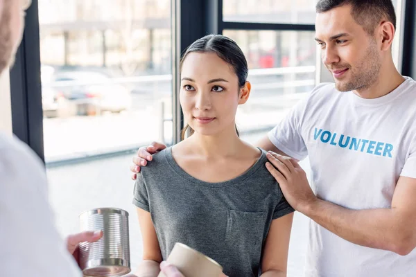 Handsome volunteer hugging asian woman taking canned food in charity center — Stock Photo