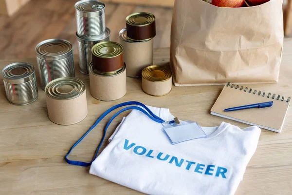 T-shirt branca com inscrição voluntária, crachá, latas, saco de papel e caderno em mesa de madeira — Fotografia de Stock