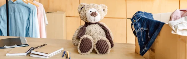 Panoramic shot of wooden table with teddy bear, carton box with clothes and notebooks — Stock Photo