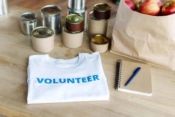 White t-shirt with blue volunteer inscription, tins, paper bag with apples and notebook on wooden table — Stock Photo