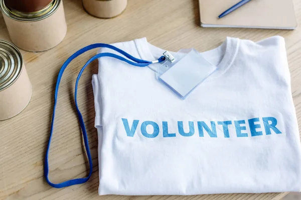 Camiseta blanca con inscripción de voluntario azul, insignia y latas en la mesa de madera - foto de stock