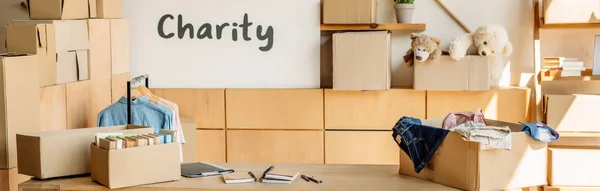 Panoramic shot of cardboard boxes with books and clothes, wooden cabinets and placard with charity inscription — Stock Photo