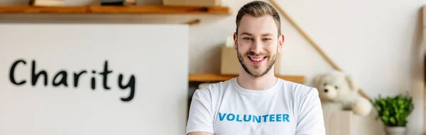 Panoramic shot of handsome, smiling volunteer standing near placard with charity inscription — Stock Photo
