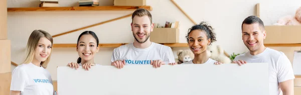 Panoramic shot of cheerful multicultural volunteers holding empty placard and looking at camera — Stock Photo