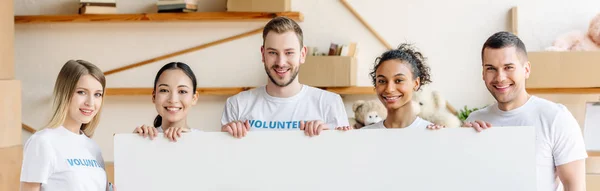 Plano panorámico de alegres voluntarios multiculturales sosteniendo un cartel vacío y mirando a la cámara - foto de stock