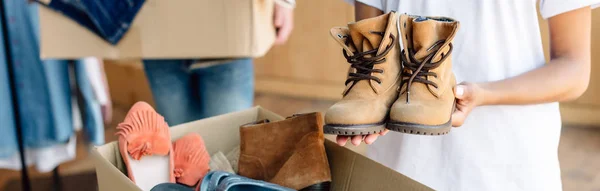 Panoramic shot of african american volunteer holding kids shoes while standing near carton box with footwear — Stock Photo