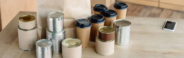 Plano panorámico de latas, vasos desechables, bolsa de papel y teléfono inteligente en mesa de madera - foto de stock