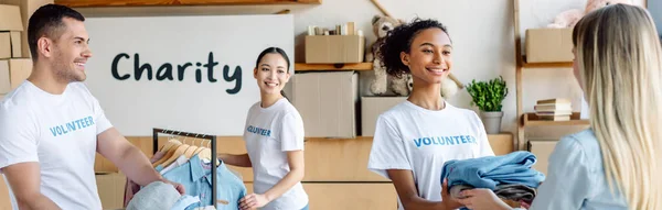 Panoramic shot of smiling multicultural volunteers giving clothes to woman — Stock Photo