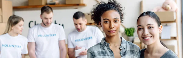 Panoramic shot of pretty multicultural women looking at camera while standing near volunteers in charity center — Stock Photo