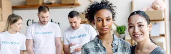 Foto panoramica di donne abbastanza multiculturali guardando la fotocamera mentre in piedi vicino ai volontari nel centro di beneficenza — Foto stock