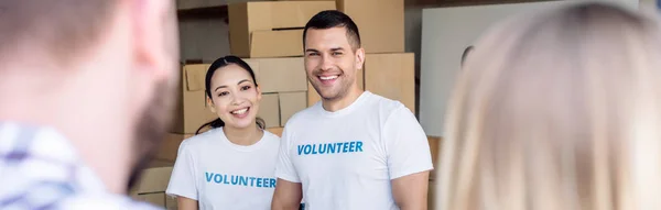 Панорамный снимок улыбающихся мультикультурных волонтеров, встречающих людей в благотворительном центре — стоковое фото