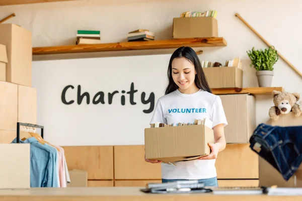 Jolie bénévole asiatique tenant boîte en carton tout en se tenant près de la plaque avec inscription de charité — Photo de stock