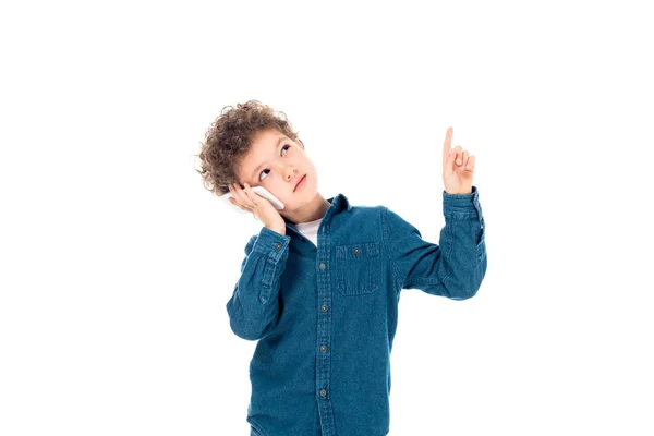 Niño rizado pensativo en camisa de mezclilla hablando en el teléfono inteligente y mostrando idea signo aislado en blanco - foto de stock
