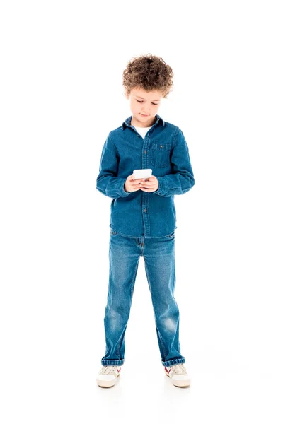 Полный вид концентрированного ребенка в джинсовой одежде с помощью смартфона, изолированного на белом — стоковое фото