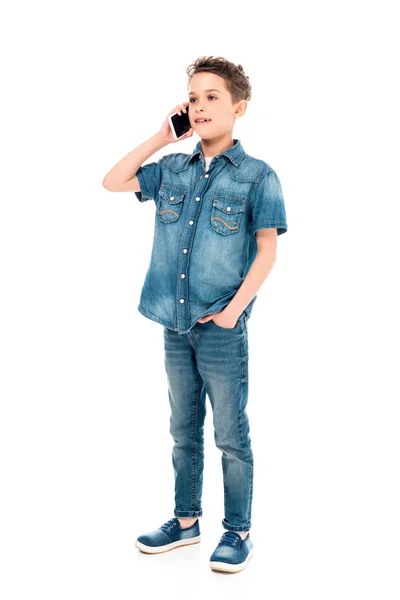 Vista completa del niño de pie con la mano en el bolsillo y hablando en el teléfono inteligente aislado en blanco - foto de stock