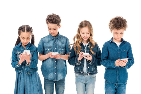 Cuatro niños en ropa de mezclilla usando teléfonos inteligentes aislados en blanco - foto de stock