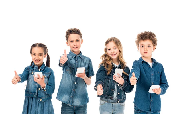 Cuatro niños en ropa de mezclilla usando teléfonos inteligentes y mostrando los pulgares hacia arriba aislados en blanco - foto de stock