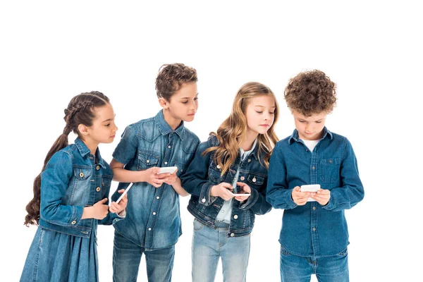 Cuatro niños en ropa de mezclilla usando teléfonos inteligentes aislados en blanco - foto de stock