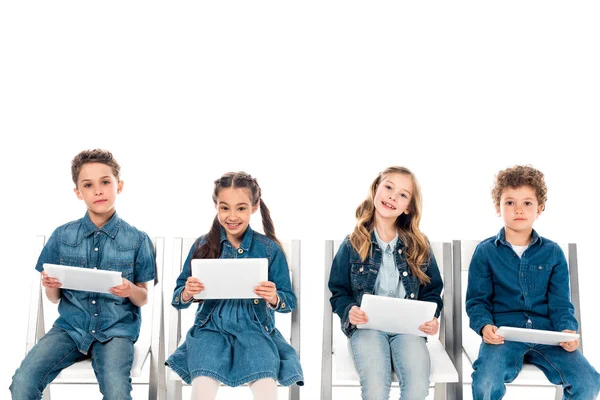 Четыре улыбающихся ребенка в джинсовой одежде сидит на стульях и с помощью цифровых таблеток изолированы на белом — стоковое фото