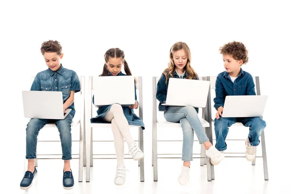 Cuatro niños en ropa de mezclilla sentados en sillas y el uso de computadoras portátiles en blanco - foto de stock