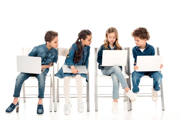 Cuatro niños en ropa de mezclilla sentados en sillas y el uso de computadoras portátiles en blanco - foto de stock