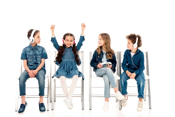 Four kids in denim clothes sitting on chairs and listening music in headphones on white — Stock Photo