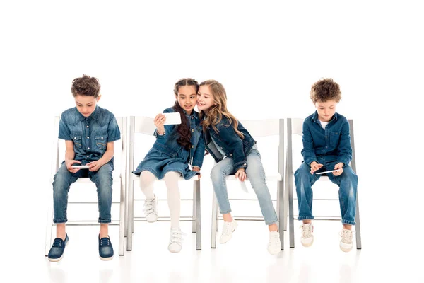 Niños en ropa de mezclilla sentados en sillas y tomando selfie en blanco - foto de stock