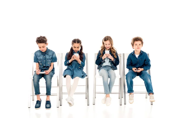 Cuatro niños en ropa de mezclilla sentados en sillas y usando teléfonos inteligentes en blanco - foto de stock