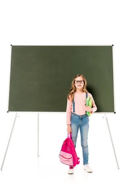 Vista completa de la colegiala en gafas que sostienen la mochila y libros cerca de pizarra aislada en blanco - foto de stock