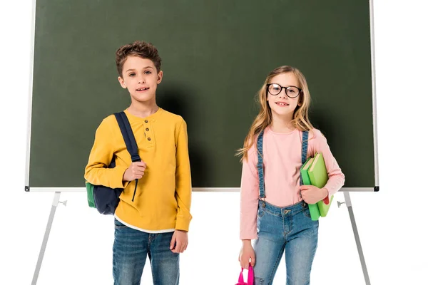 Two schoolchildren with backpacks and books standing near blackboard isolated on white — Stock Photo