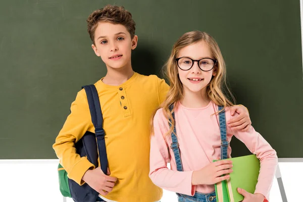 Duas crianças em idade escolar com mochilas e livros em pé perto de quadro negro isolado em branco — Fotografia de Stock