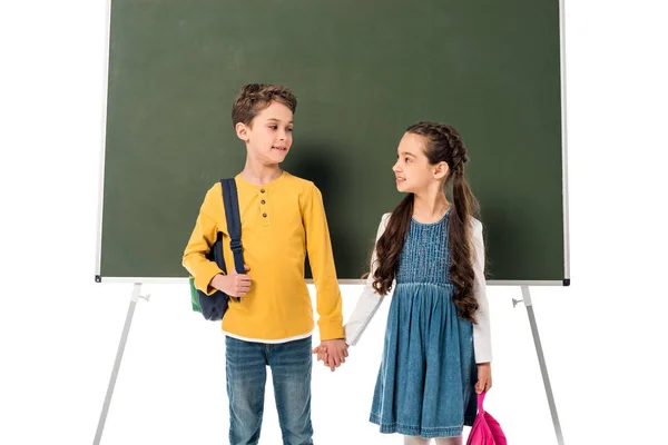 Duas crianças em idade escolar com mochilas de mãos dadas perto de quadro-negro isolado em branco — Fotografia de Stock