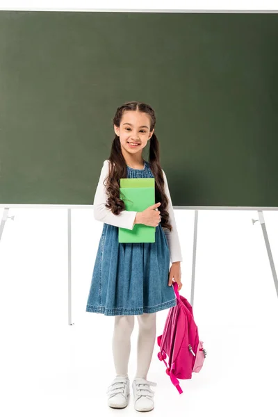 Vue pleine longueur de l'écolière souriante avec sac à dos et livres debout près du tableau noir isolé sur blanc — Photo de stock