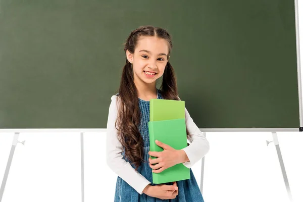 Colegiala sonriente sosteniendo libros cerca de pizarra aislado en blanco - foto de stock