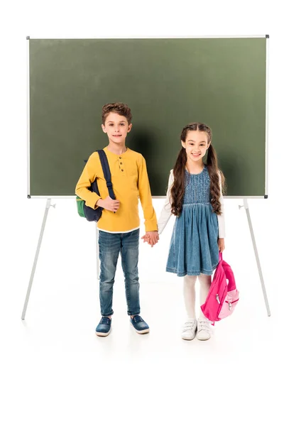 Visão de comprimento total de duas crianças em idade escolar com mochilas de mãos dadas perto de quadro negro isolado em branco — Fotografia de Stock