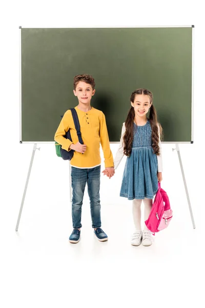 Vista completa de dos escolares con mochilas cogidas de la mano cerca de pizarra aislada en blanco - foto de stock