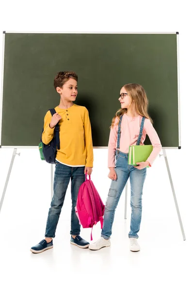 Ganzkörperansicht von zwei Schulkindern mit Rucksäcken, die sich in der Nähe der Tafel anschauen, isoliert auf weißem Grund — Stockfoto