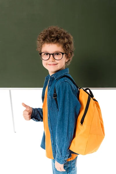 Vista de alto ângulo de estudante sorrindo em óculos de pé perto de quadro-negro e mostrando polegar para cima isolado em branco — Fotografia de Stock