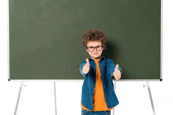 Écolier souriant dans des lunettes debout près du tableau noir et montrant pouces isolés sur blanc — Photo de stock