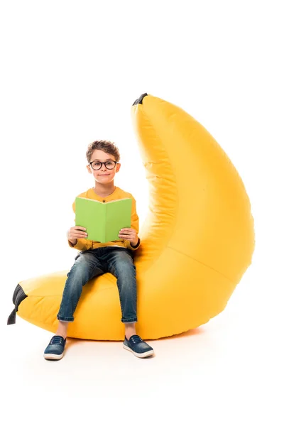 Kind in lässiger Kleidung sitzt auf Bohnensackstuhl und liest Buch auf weiß — Stockfoto