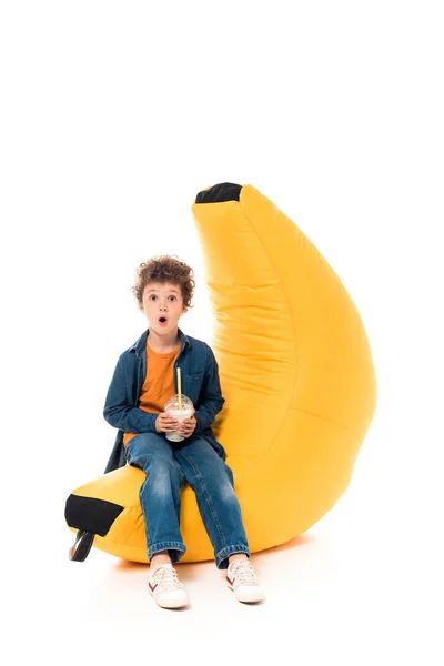 Niño sorprendido en jeans sentados en la silla de la bolsa de frijol y sosteniendo el batido en blanco - foto de stock