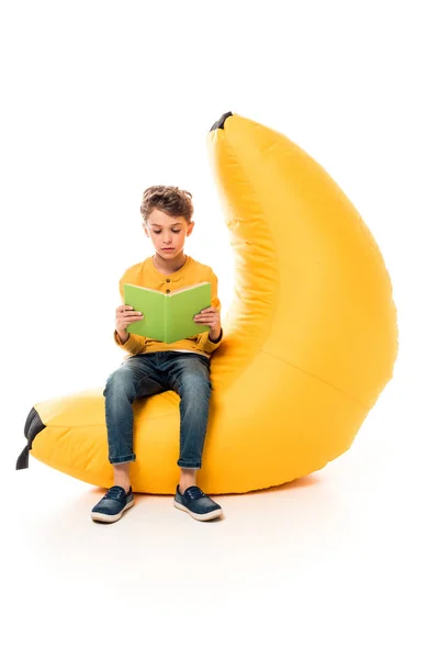 Kind in lässiger Kleidung sitzt auf Bohnensackstuhl und liest Buch auf weiß — Stockfoto