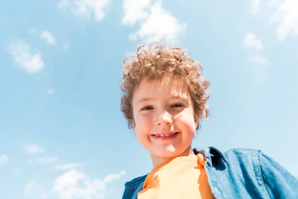 Vista angolo basso del bambino riccio sorridente sotto il cielo blu — Foto stock