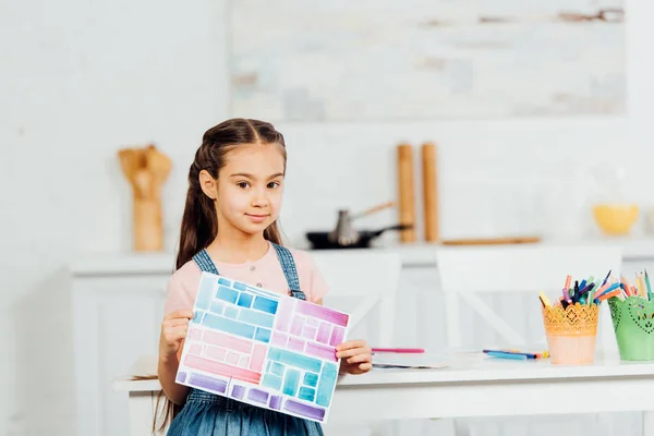 Niedliches Kind, das Papier mit bunten Streifen hält, während es zu Hause am Tisch steht — Stockfoto