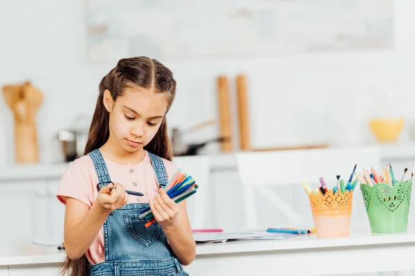 Niedliches Kind hält bunte Filzstifte und Stifte in der Hand, während es zu Hause am Tisch steht — Stockfoto