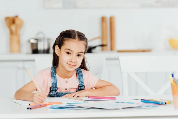 Niedliches Kind schaut in die Kamera, während es zu Hause Farbstift in der Nähe von Papier hält — Stockfoto