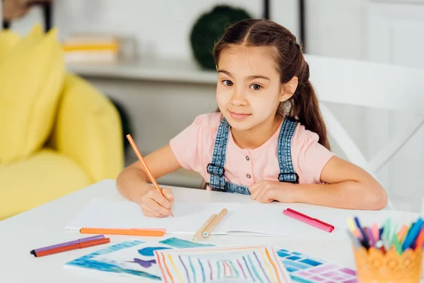 Messa a fuoco selettiva del bambino felice guardando la fotocamera tenendo la matita di colore vicino alla carta a casa — Foto stock