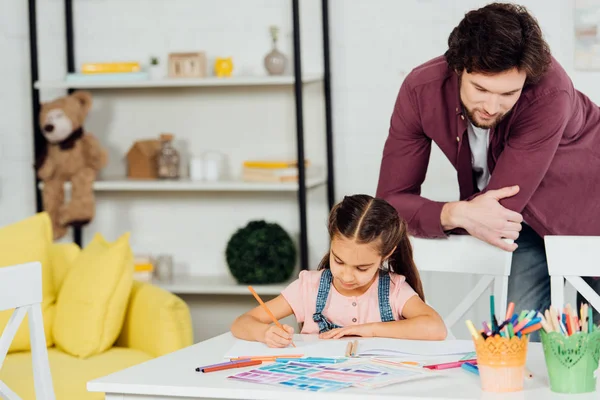 Schöner Vater steht neben süßer Tochter, die zu Hause zeichnet — Stockfoto