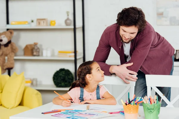 Hermoso padre de pie cerca de linda hija sosteniendo lápiz de color en casa - foto de stock