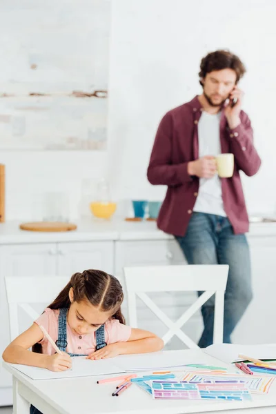 Messa a fuoco selettiva del bambino carino disegno su carta vicino al padre parlando su smartphone e tenendo in mano tazza — Foto stock